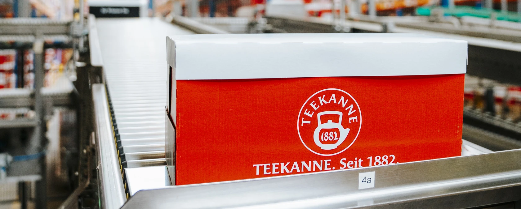 Eine rote Teekanne mit der Aufschrift Seit 1882 bewegt sich auf einem Förderband in einer hellen Fabrik.