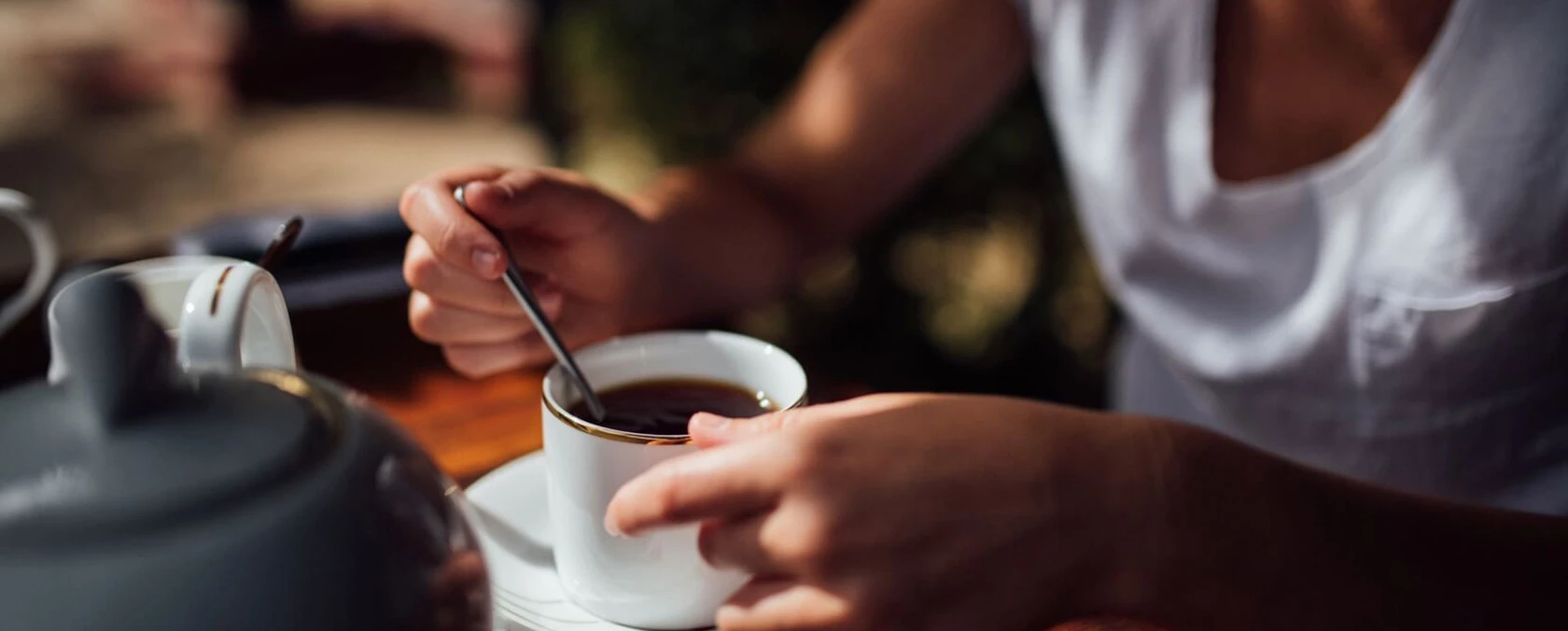 Hände rühren Kaffee in einer weißen Tasse auf einem Holztisch, Teekanne in der Nähe, Fokus auf den Händen.