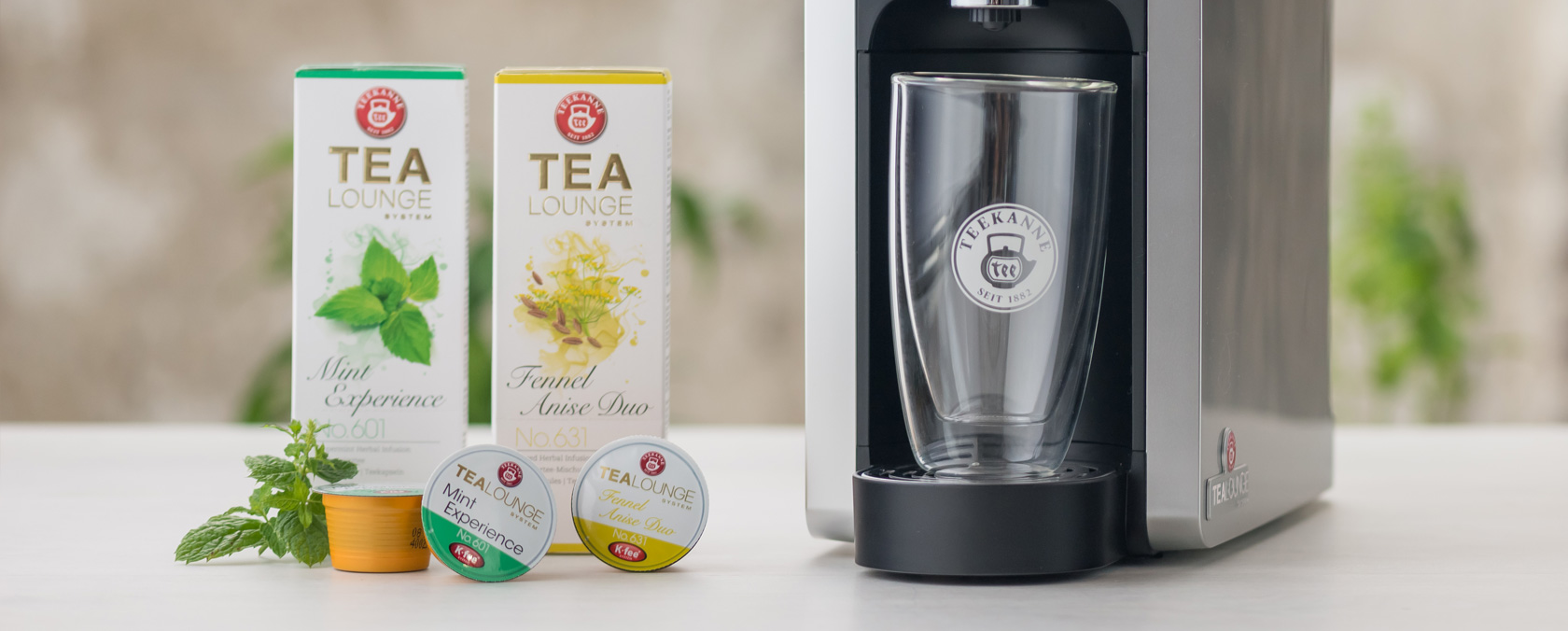 Teemaschine, Klarglas, zwei Tea Lounge System Boxen, drei verschiedene Kapseln auf hellem Hintergrund.