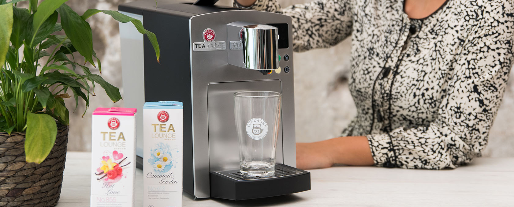 Person brüht Tee mit Maschine, Glas unter Spender, bunte Teedosen und Pflanze in der Nähe.