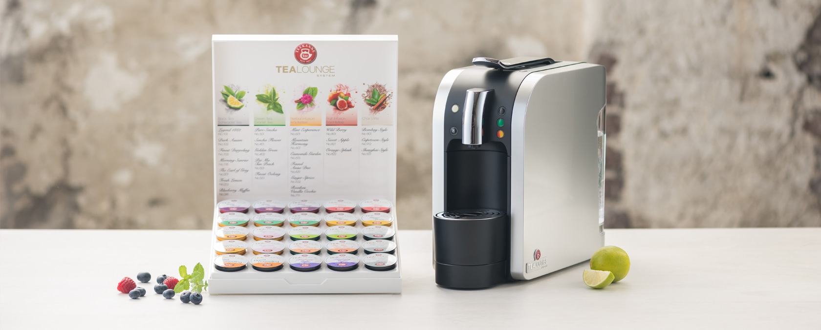 Teeaufbrühmaschine mit verschiedenen Geschmackskapseln, frischen Beeren und Limette auf einer weißen Oberfläche.