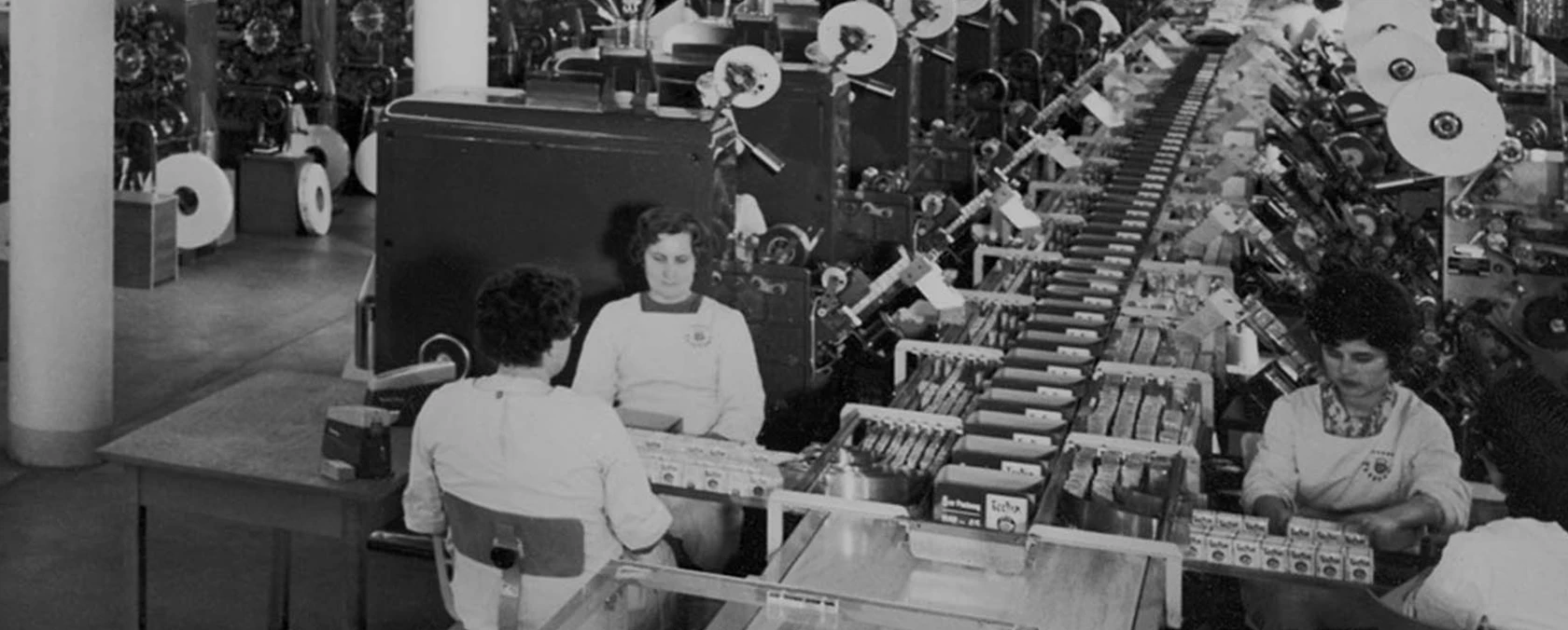 Frauen arbeiten an einem Fließband in einer geschäftigen Fabrik voller Maschinen und Verpackungsmaterial.