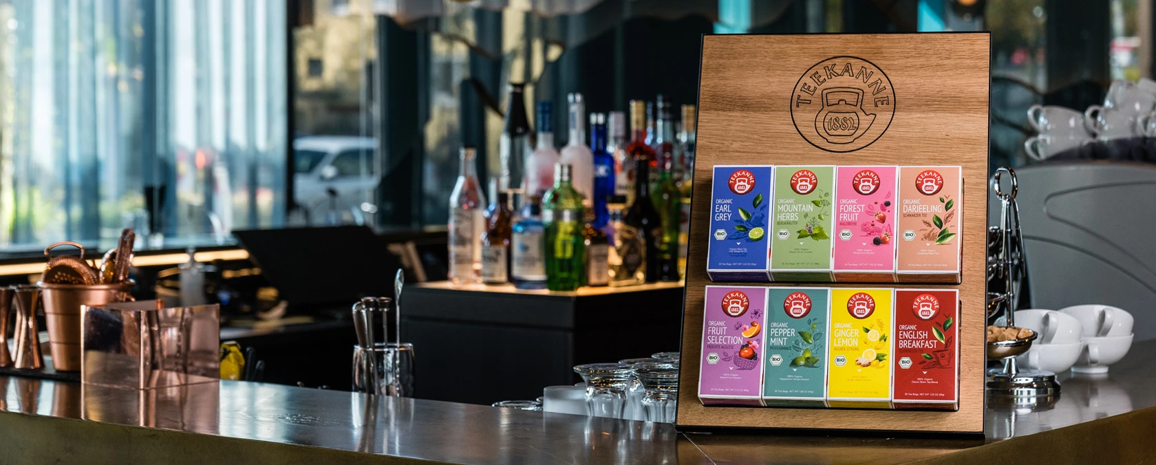 Cafétheke mit verschiedenen Bio-Teekisten, Gläsern, Kaffeezubehör und Sonnenlicht aus großen Fenstern.