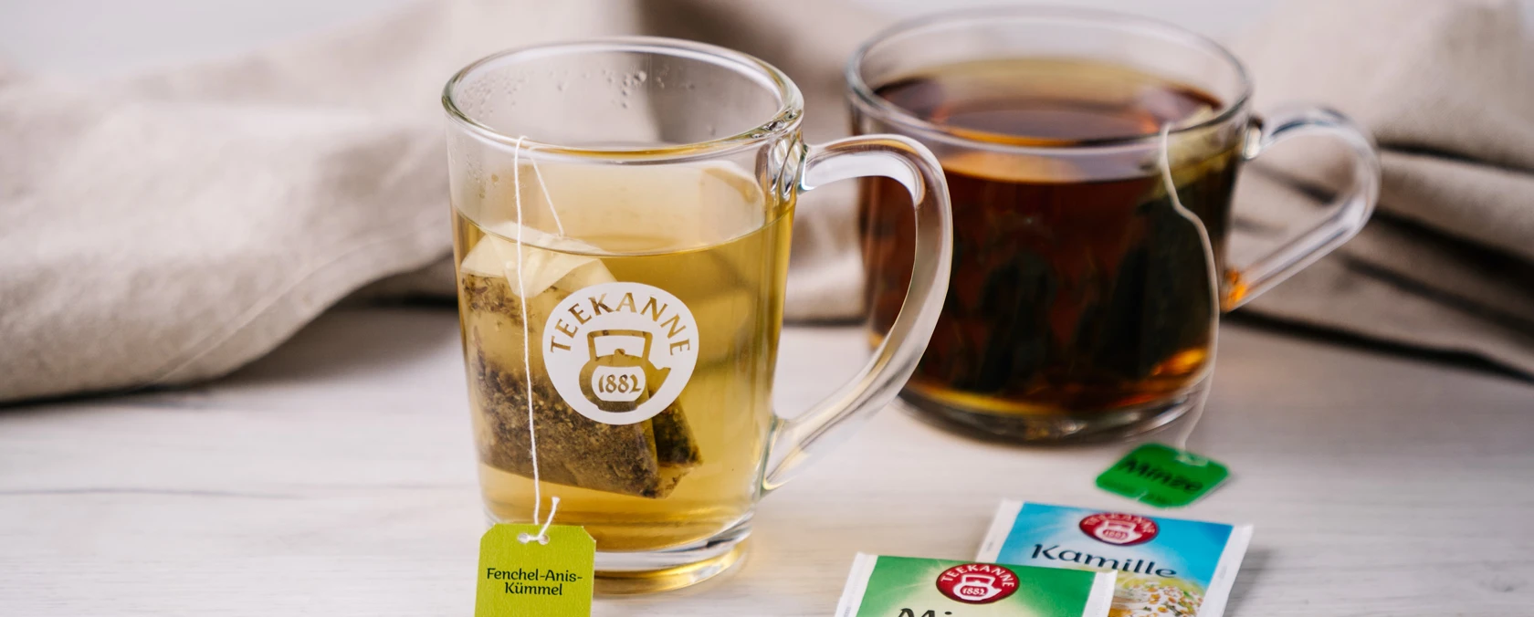 Zwei Tassen Tee, eine hellgelbe und eine dunkle, mit Kräuterteepäckchen daneben.