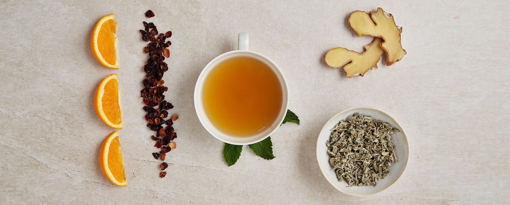Teetasse mit Orangenscheiben, getrockneten Beeren, Minze, Ingwer und Kräutern auf hellem Hintergrund.