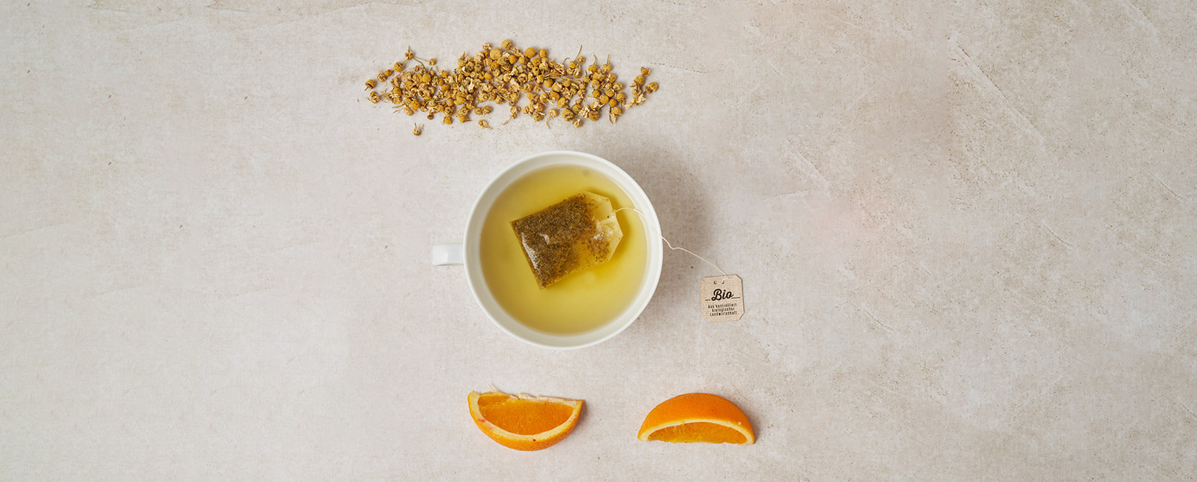 Weiße Tasse Kamillentee mit TAZO-Anhänger, oben lose Blüten, unten Orangenscheiben.