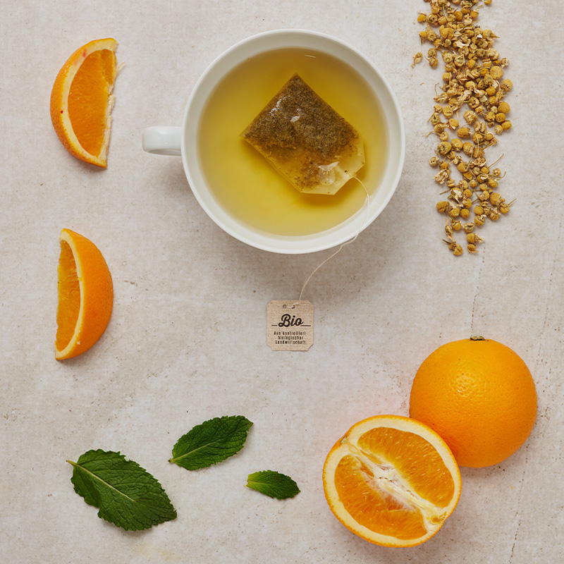 Weiße Tasse Kräutertee mit Teebeutel, Orangenscheiben, ganzer Orange, getrockneten Kräutern und frischer Minze.
