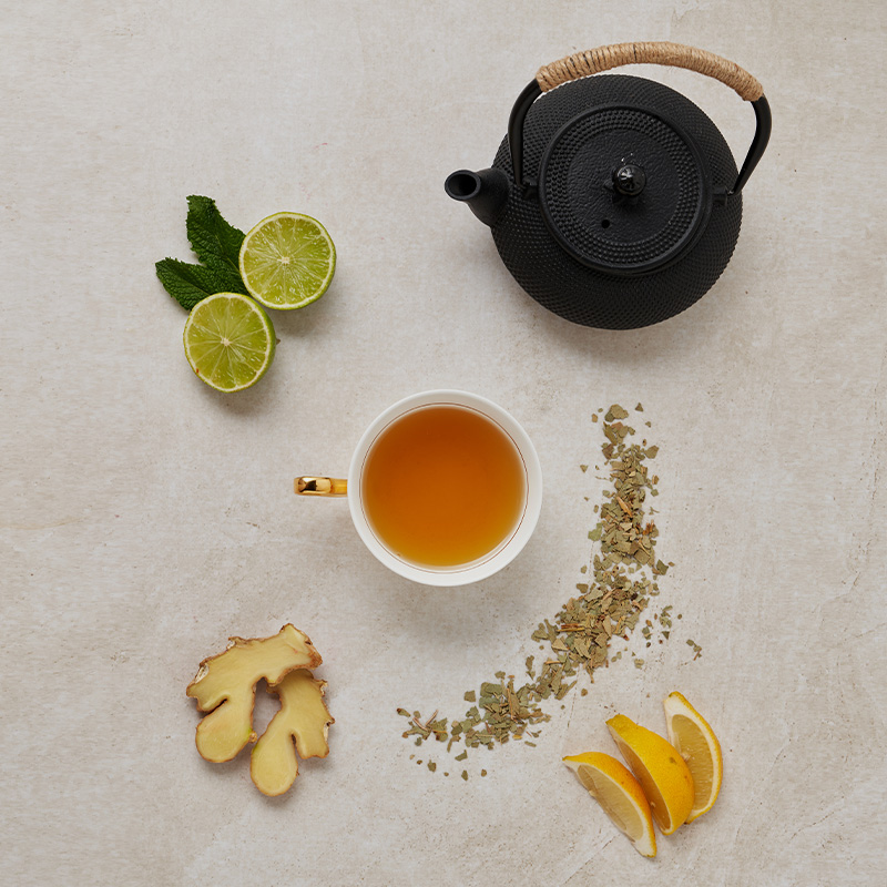 Tasse Tee mit Teekanne, Limette, Minze, Ingwer, losen Teeblättern und Orangenspalten arrangiert.