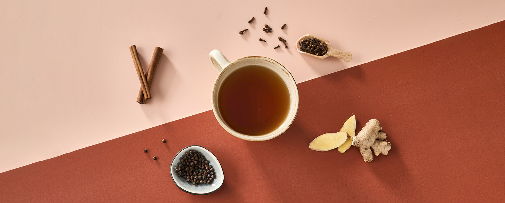 Tasse Tee mit Zimt, Pfefferkörner, Nelken, Ingwer und Holzschaufel auf rosa Hintergrund.