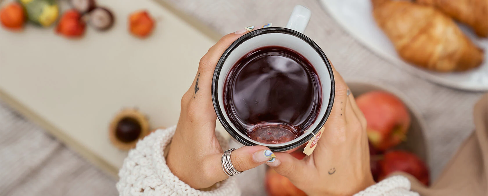 Hand mit Ringen und lackierten Nägeln, die einen Teebecher, einen Picknickaufstrich mit Obst, Croissant und Snacks hält.