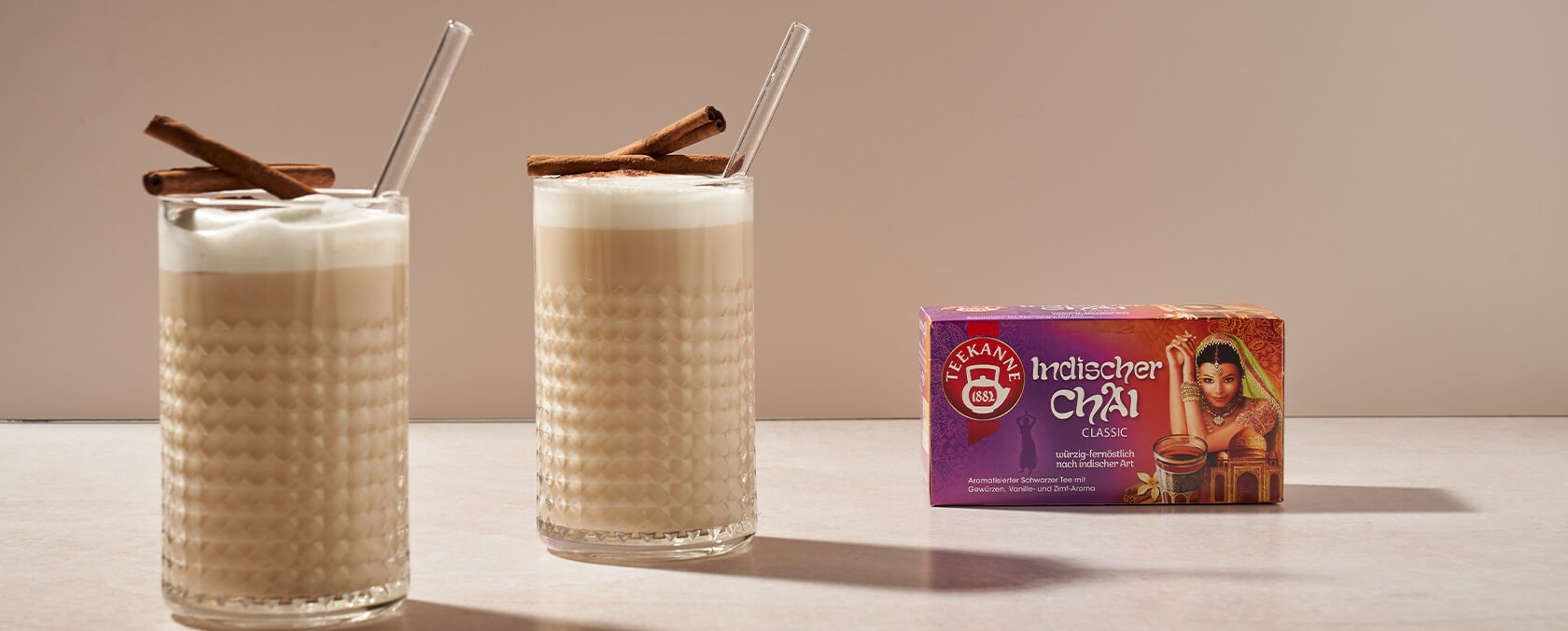Zwei schaumige Chai-Gläser mit Zimtstangen neben einer bunten Indischer Chai Classic Teedose.