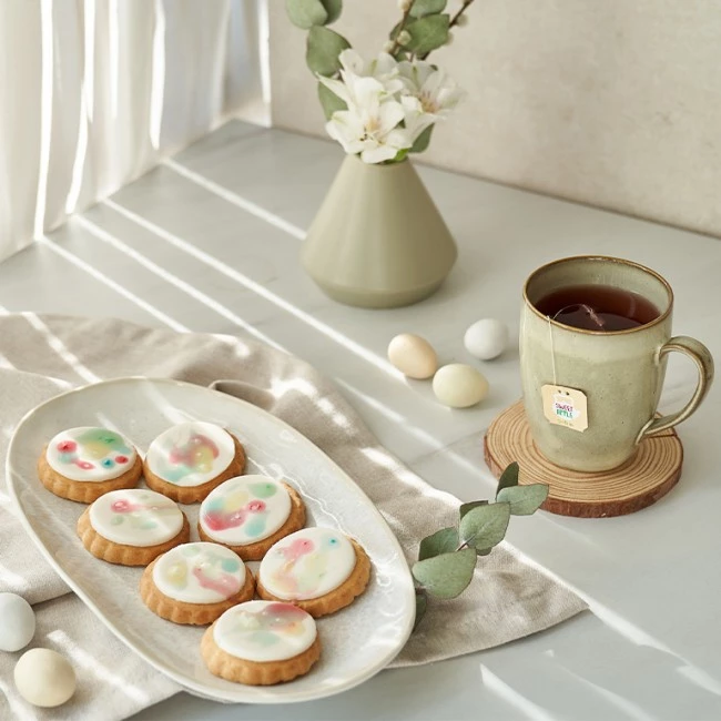 Ovale Kekse mit Zuckerguss und eine Tasse Tee neben pastellfarbenen Eiern und weißen Blumen im Sonnenlicht.