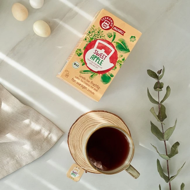 Tasse süßer Apfeltee, Teedose, drei Eier, beiges Tuch, Eukalyptuszweig auf heller Oberfläche.