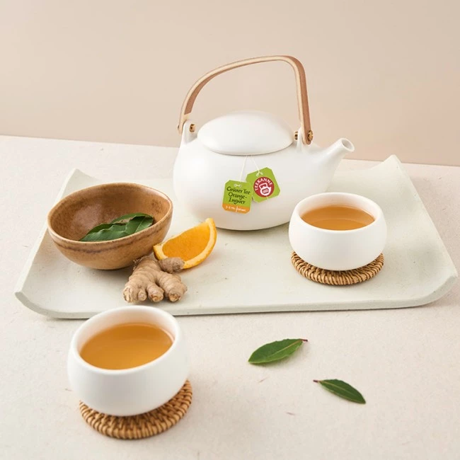 Teekanne, Tassen Tee, Ingwerwurzel, Orangenscheibe und grüne Blätter auf einem Tablett angeordnet.