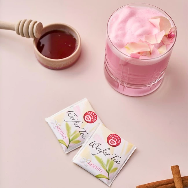 Rosa Tee mit Rosenblättern, Honiglöffel in Honigschale, Jasminteebeutel, Zimtstange.