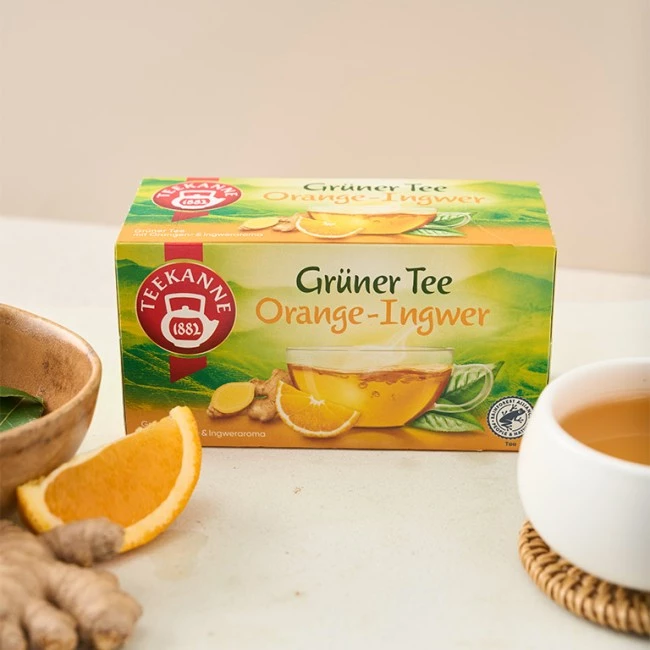 Teekanne Grüner Tee Orange-Ingwer-Box mit Tee, Orange, Ingwer und Teetasse auf dem Tisch.