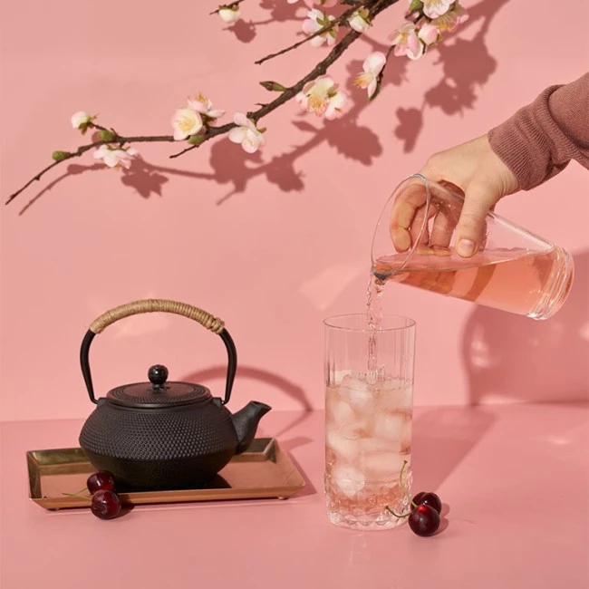 Hand gießt rosa Eistee in ein Glas mit Eis; Kirschen, Teekanne und Kirschblüten in der Nähe.
