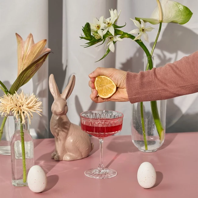 Hand presst Zitrone über rotes Getränk; Keramikhase, Eier, Blumen und weiße Vorhänge im Hintergrund.