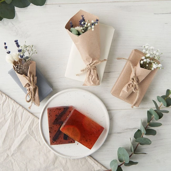 Drei dekorative Seifen mit getrockneten Blumen, zwei rote durchscheinende Seifen auf einem Teller, Grünpflanzen und beigefarbenem Stoff.