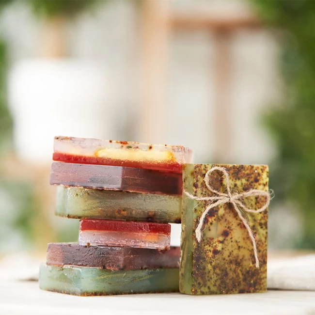 Stapel bunter handgemachter Seifen, ein grüner Riegel mit Kräutern und einer Schleife aus Bindfäden, verschwommener Blätterhintergrund.
