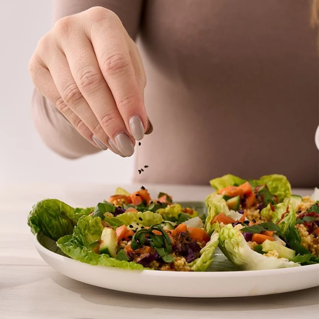 Eine Hand streut Samen über Salatwraps mit leuchtendem Gemüse auf einem weißen Teller vor hellem Tischhintergrund.