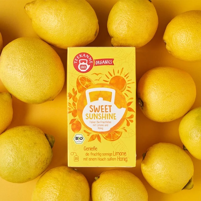 Teekanne Sweet Sunshine Teebox mit Zitronen-, Honig- und Sonnenillustrationen, umgeben von frischen Zitronen.