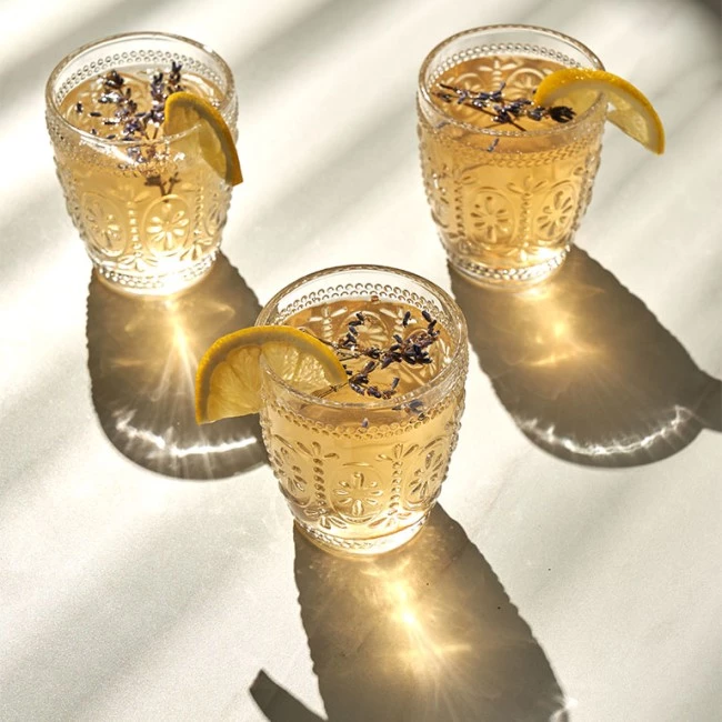 Drei dekorierte Gläser mit Zitronen-Lavendel-Getränken werfen Schatten und Spiegelungen auf eine sonnenbeschienene Oberfläche.