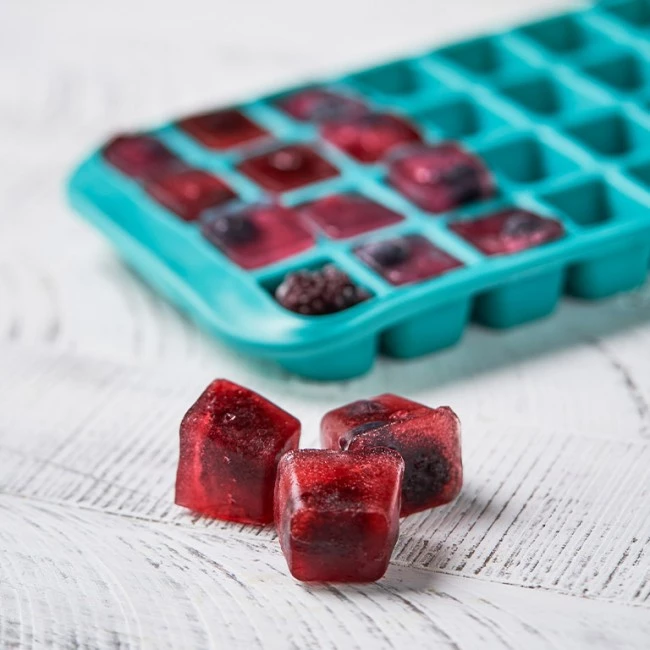 Drei mit roten Beeren gefüllte Eiswürfel auf weißem Holz, dahinter ein blaugrünes Tablett mit ähnlichen Würfeln.