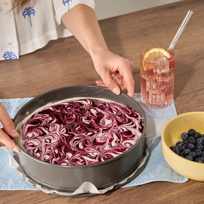 Person stellt marmorierten Beerenkäsekuchen neben Blaubeeren und rosa Getränk mit Zitronenscheibe auf den Tisch.