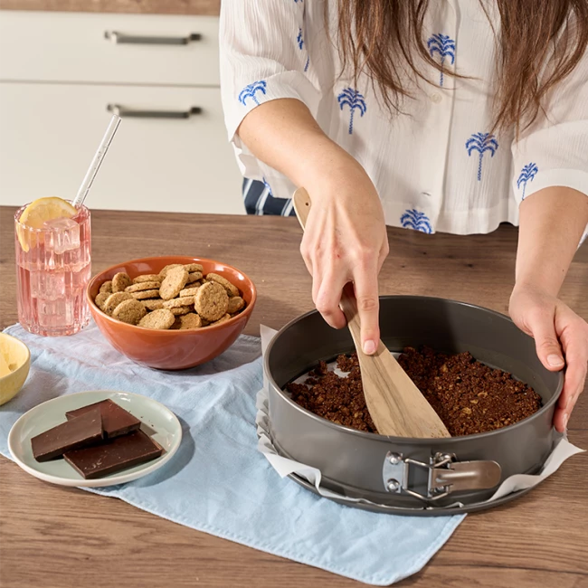 Frau drückt Krümelboden in Springform; Kekse, Schokolade und rosa Getränk auf der Theke.