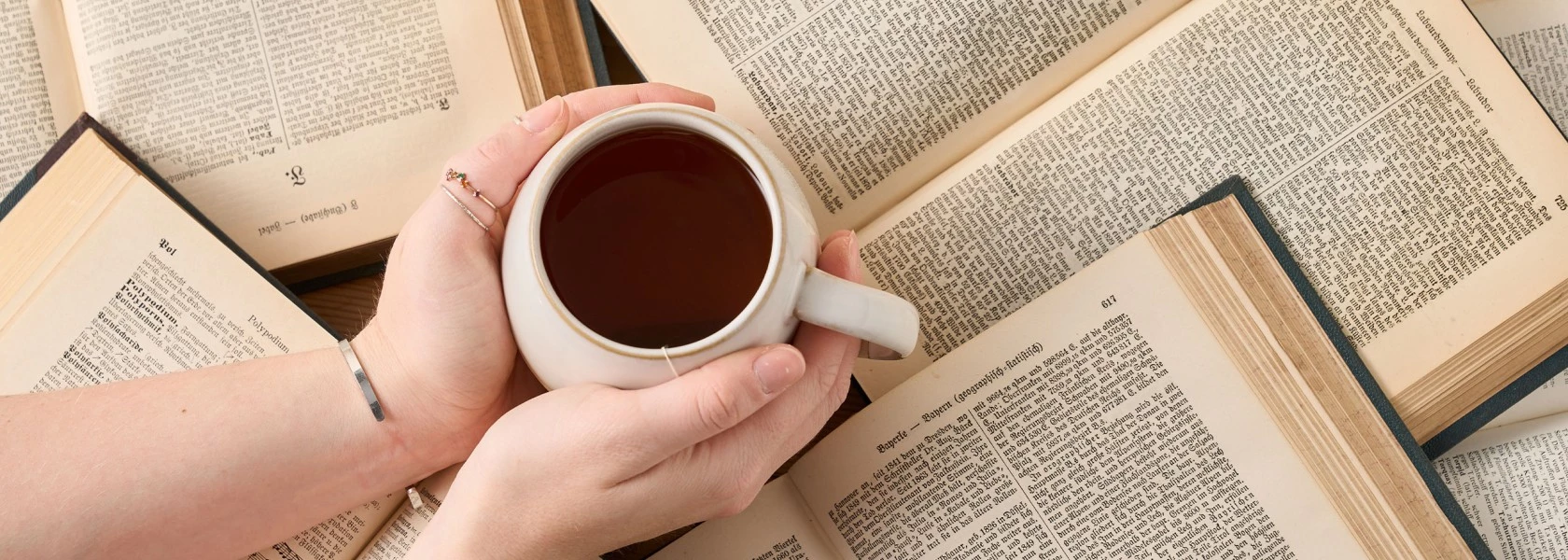 Person hält weiße Kaffeetasse über einem Tisch mit offenen Büchern, von oben betrachtet.
