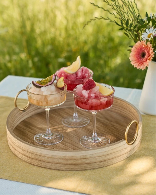 Drei farbenfrohe gefrorene Cocktails mit Fruchtgarnitur auf einem Holztablett, gelbe Tischdecke, Blumen und Grünpflanzen dahinter.