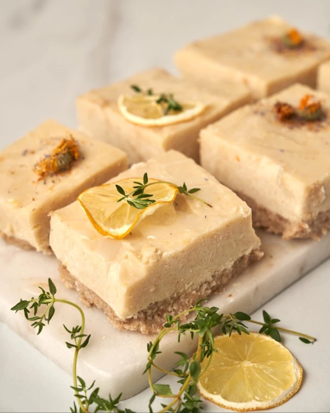 Sechs Zitronenschnitten mit Streuselboden, Zitronenscheiben und Kräutern auf Marmorplatte, dekorativ garniert.