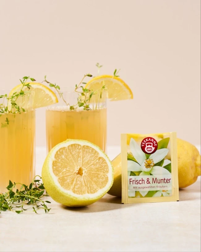 Zwei Gläser Kräutertee mit Zitronen, frischen Kräutern und einem gelben „Frisch & Munter“-Teepäckchen.