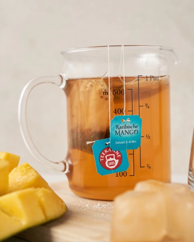 Glasmessbecher mit Mango-Teebeuteln, ziehender Tee neben Eiswürfeln und frischen Mangoscheiben.