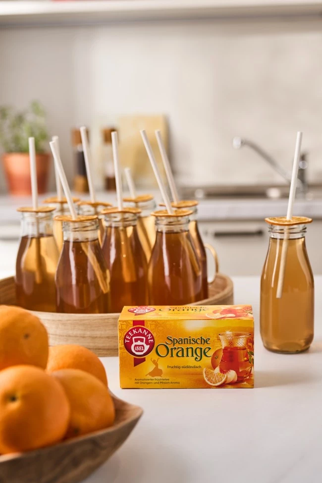 Sechs Orangengetränke mit Strohhalmen auf Tablett; Teebox „Spanische Orange“ und Orangen im Vordergrund.