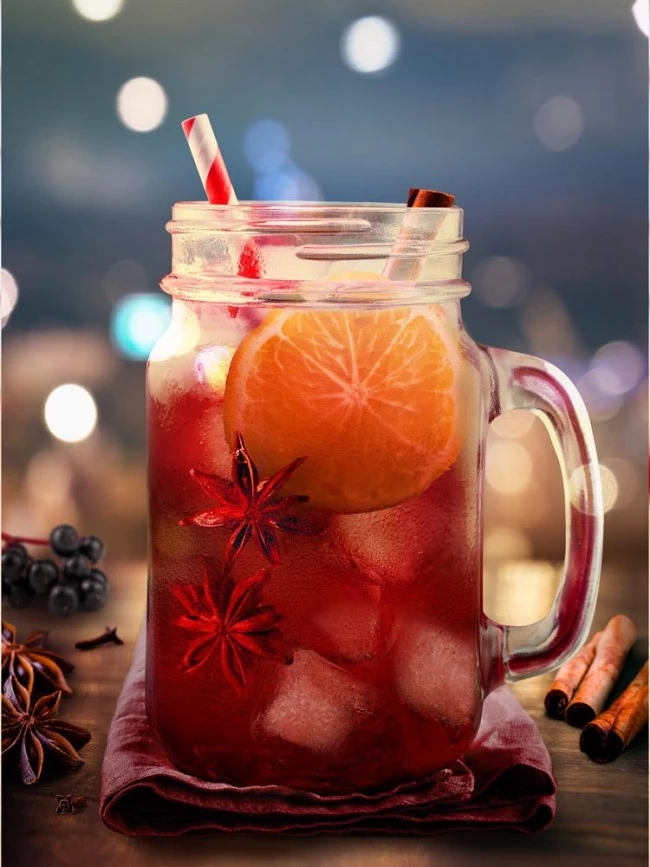 Einmachglas mit eisgekühltem roten Getränk mit Orange, Sternanis, Zimt, gestreiftem Stroh; daneben Gewürze.