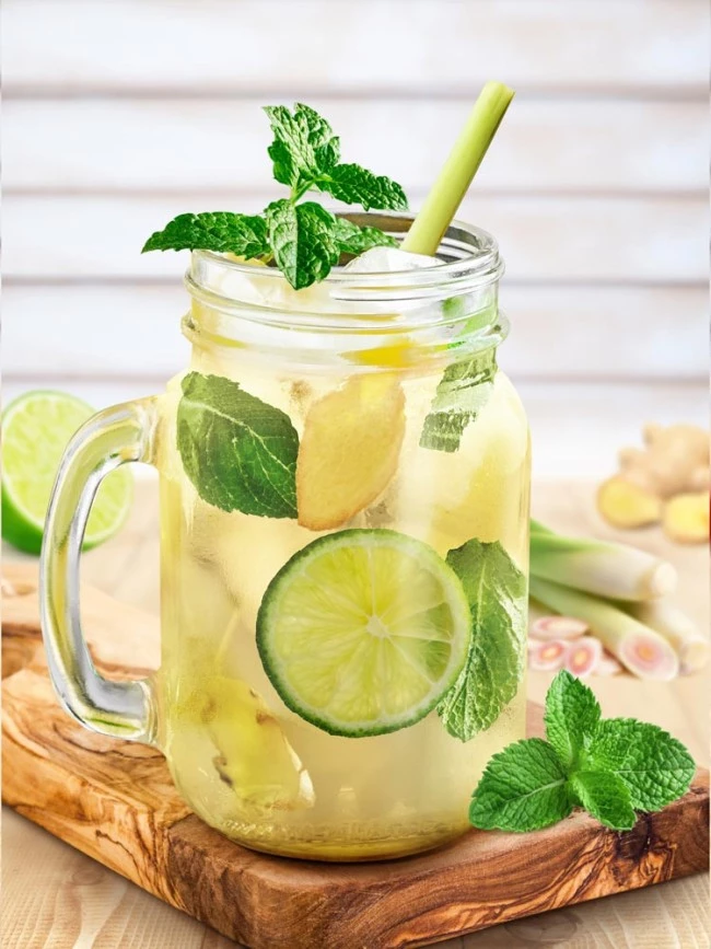 Einmachglas mit Limetten-, Ingwer-, Minz- und Zitronengrasgetränk auf einem Holzbrett, umgeben von frischen Zutaten.
