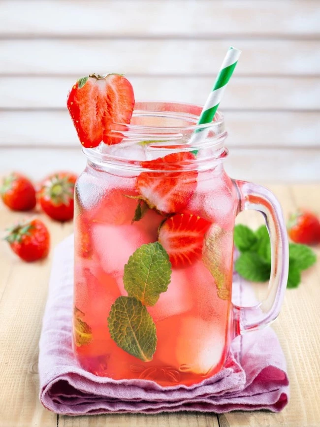 Einmachglas mit rosa Erdbeergetränk mit Minze, Eis und Strohhalm auf rosa Tuch; dahinter Erdbeeren.