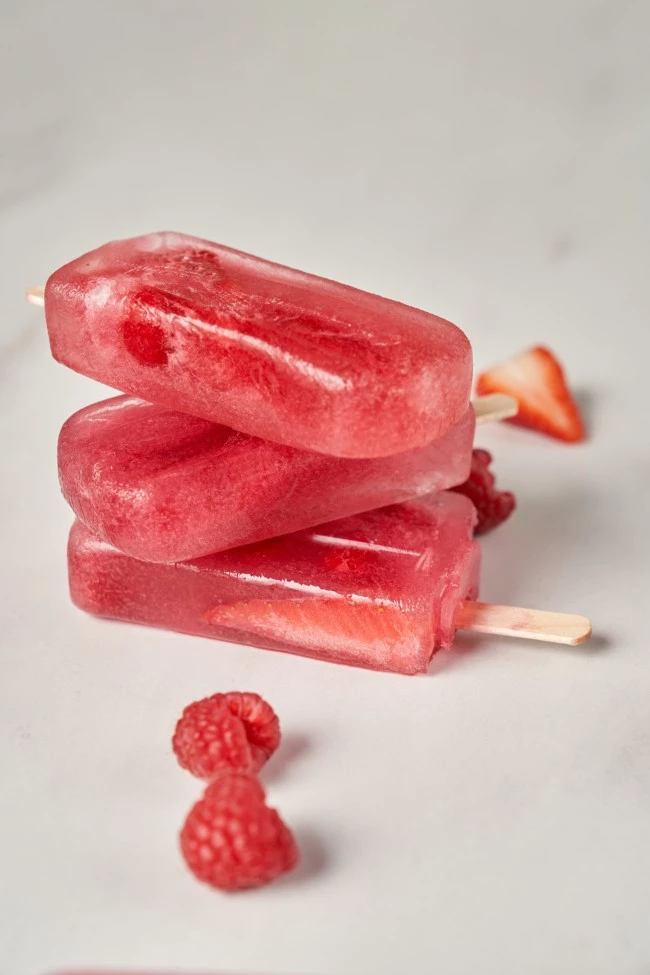 Drei rote Fruchteis am Stiel, gestapelt mit Himbeeren und einer Erdbeere auf einer weißen Oberfläche, ein Sommergenuss.