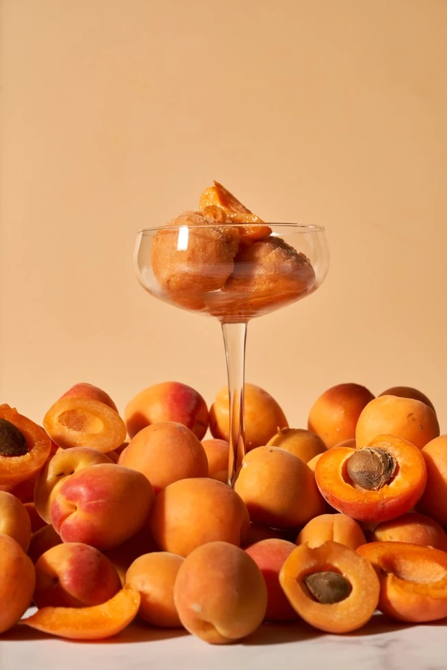 Aprikoensorbet im Glasbecher, garniert mit frischen Aprikosen, umgeben von ganzen und halbierten Aprikosen.