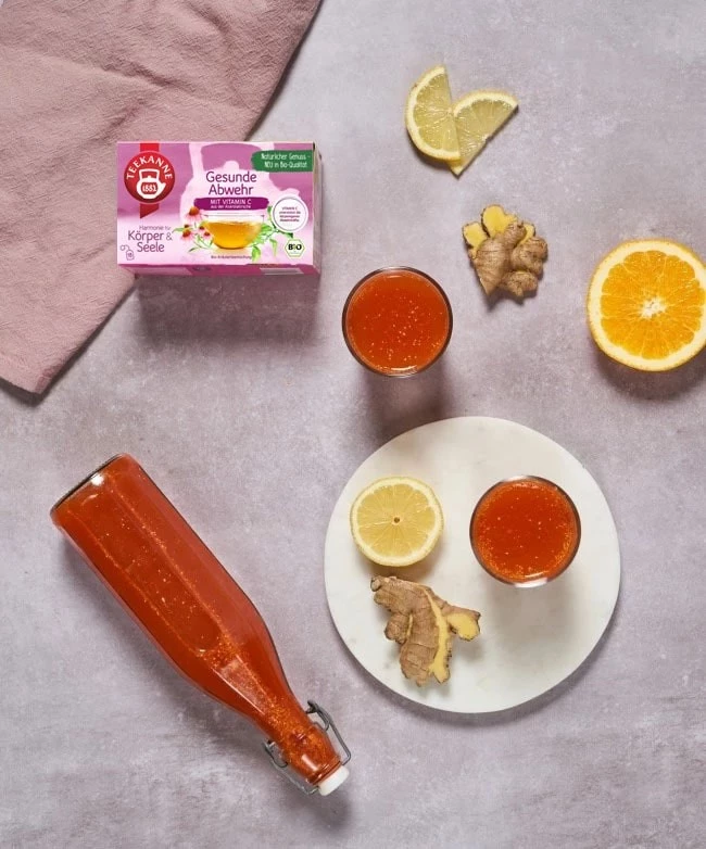 Teekanne Gesunde Abwehr auf einem Tisch mit einem Vitamin Shot, dekoriert mit Ingwer, Zitronen- und Orangenscheiben.