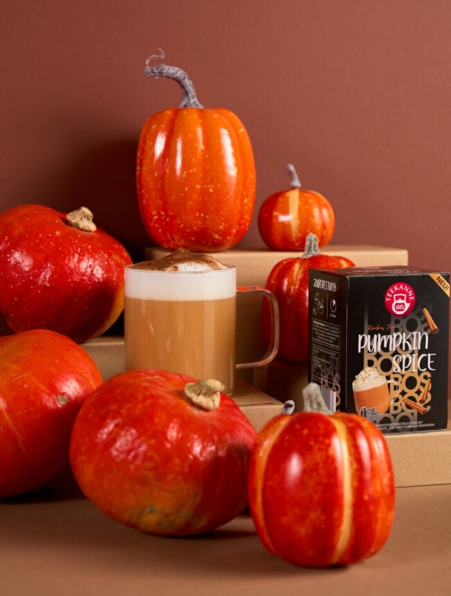 Pumpkin Spice Latte in einem Glasbecher mit Kürbissen und Packung "TEEKANNE Pumpkin Spice" Tee auf einem braunen Hintergrund.