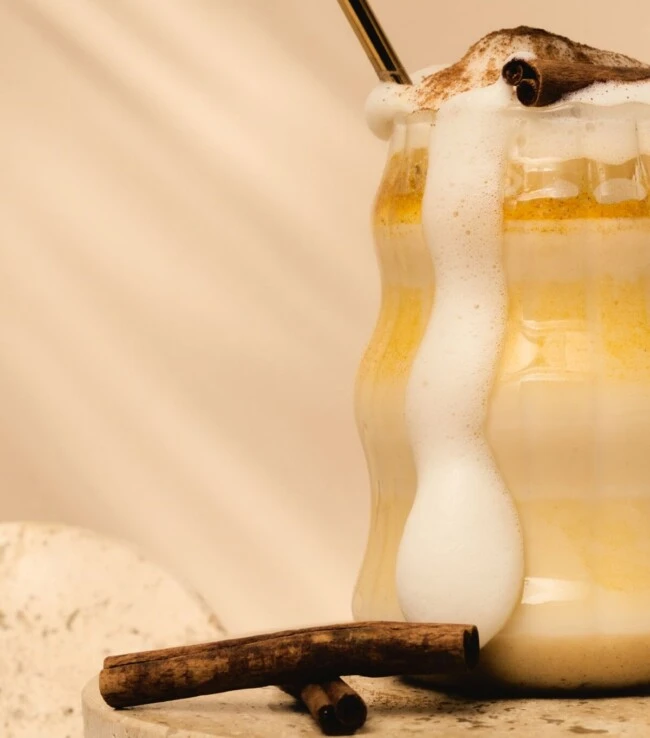 Schaumiges, cremiges Getränk mit überquellendem Schaum, gekrönt von einer Zimtstange; Strohhalm und Löffel oben; Zimtstangen daneben.