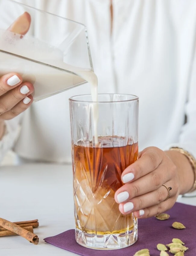 Milch aus dem Krug in Eistee gegossen; weißer Nagellack, Gewürze auf lila Serviette neben dem Glas.