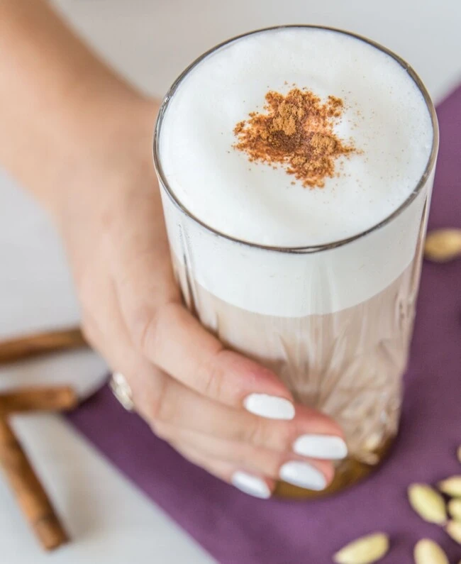 Eine Hand mit weißen Nägeln hält einen schaumigen Chai Latte; dahinter Zimtstangen und Kardamom auf einer lila Serviette.