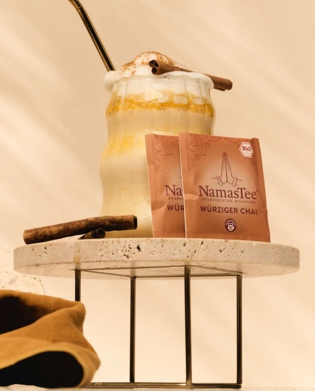 Geschichteter Chai mit aufgeschäumter Milch und Zimt, Metallstrohhalm, Teepäckchen und Zimtstange auf Marmortablett.