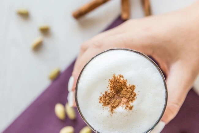 Hand mit schaumiger Milch mit Zimt, Kardamomschoten und Zimtstangen auf lila Serviette unten.