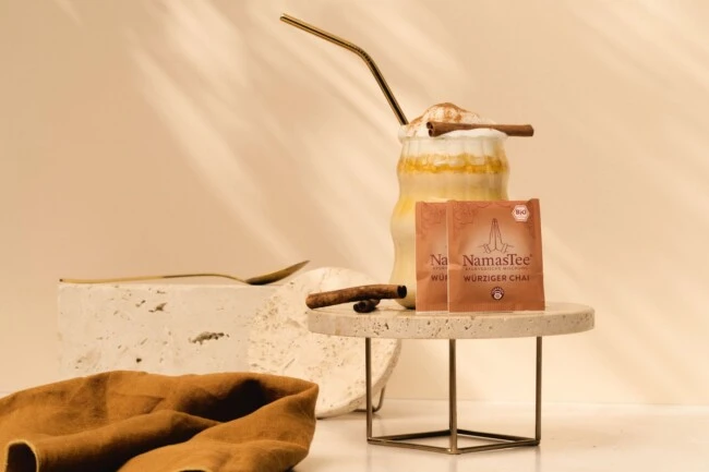 Chai mit Schlagsahne und Zimt auf Steinständer, Teepackung, zusätzliche Zimtstange und braunes Tuch daneben.