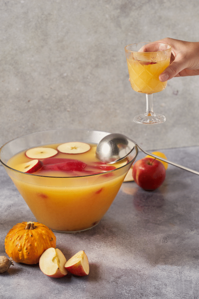Hand hält ein Glas Apfelpunsch über einer Schüssel, umgeben von Äpfeln und einem Kürbis auf grauer Oberfläche.
